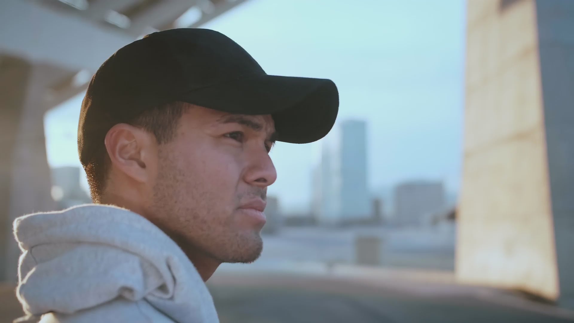 Athletic young man in a dark baseball cap and light gray hoodie under an overpass looking across a river at a city skyline. 